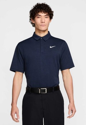 Mørkeblå polo skjorte laget av et teksturert stoff, med en knappestolpe med to knapper og en hvit Nike-logo på venstre bryst. Svart belte med sølvspenne.