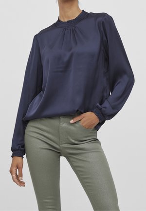 Granatowa bluzka z satyny z wysokim marszczonym kołnierzem, długimi rękawami i dopasowanymi mankietami, zestawiona z metalicznymi oliwkowozielonymi obcisłymi spodniami.