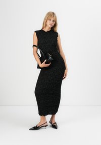 Vrouw in een textuur zwart mouwloos midi-jurk, met een zwarte handtas, staand in zwarte puntige slingback hakken op een witte achtergrond.