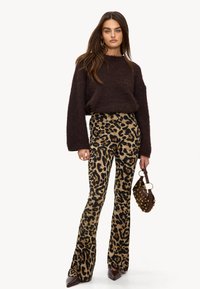 Jersey marrón de pelito, pantalones de campana con estampado de leopardo en beige y negro, sosteniendo un bolso marrón con tachuelas, zapatos puntiagudos marrones.