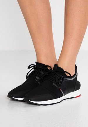 Beine, die schwarze Sportschuhe mit weißen Sohlen und schwarzen Schnürsenkeln tragen, stehen auf einem glatten, hellen Boden.