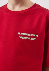 Gros plan d'une personne portant un T-shirt rouge avec le texte blanc "AMERICAN VINTAGE" sur la poitrine.