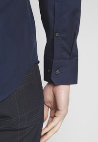 Navyblauer Hemdärmelsaum mit Knöpfen, getragen zu dunklen karierten Hosen, Hand entspannt an der Seite vor einfachem Hintergrund.