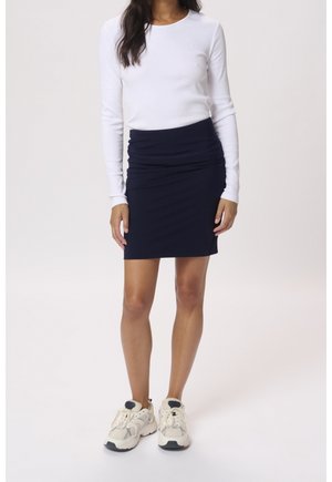 Vrouw met een wit shirt met lange mouwen, een marineblauwe geplooide kokerrok en witte robuuste sneakers, staand tegen een effen lichte achtergrond.