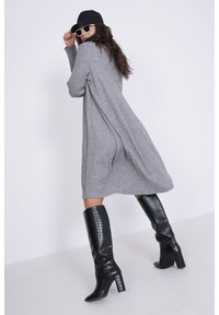 Grijze gebreide jurk met een losse pasvorm, voorzien van zijsplitten. Gepaard met glanzende zwarte kniehoge laarzen en een zwarte pet, aangevuld met witte zonnebril.