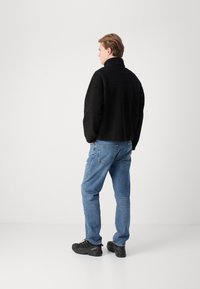 Zwarte fleecjas met hoge kraag, gedragen over lichtblauwe jeans en zwarte laarzen. De jas heeft een gestructureerd oppervlak en een ontspannen pasvorm.