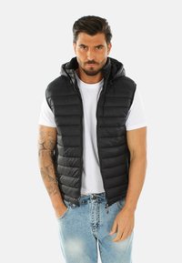 Gilet imbottito nero con cappuccio, dotato di chiusura con zip e texture a costine. Indossato su una t-shirt bianca, abbinato a jeans azzurri.