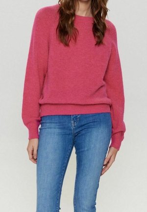Mujer con suéter rosa de manga larga y jeans ajustados azules, de pie frente a un fondo claro y liso.
