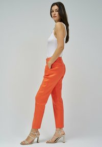 Mujer vestida con una camiseta sin mangas blanca, pantalones ajustados de color naranja brillante y sandalias de tacón beige, de pie de lado con las manos en los bolsillos.