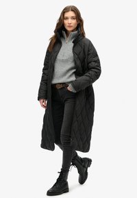 Sort quiltet lang frakke, grå striktrøje, mørke jeans og sorte snørebåndsstøvler. Bemærkelsesværdig lynlåsdetaile og høj krave på frakken.