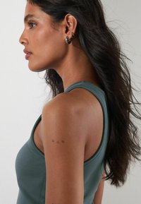 Mēteļa krāsas bez piedurknēm blūze ar apaļu dekoltē. Gluda, pieguļoša auduma. Garie, tumšie mati daļēji noseguši plecu, uz kura ir neliels ziedu tetovējums.