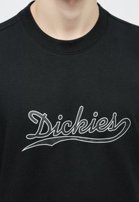 Κοντινό πλάνο ενός μαύρου πουκαμίσου με στρογγυλή λαιμόκοψη, στο στήθος του οποίου υπάρχει η λέξη "Dickies" γραμμένη με λευκά καλλιγραφικά γράμματα.