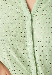 Jasnozielona koszula na guziki wykonana z teksturowanej tkaniny z małymi wycięciami i dekoltem w kształcie litery V, ozdobiona pasującymi guzikami.