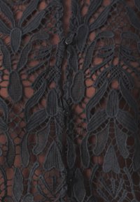 Blouse en dentelle noire avec motifs floraux et texture détaillée, au design transparent et avec de petits boutons sur le devant.