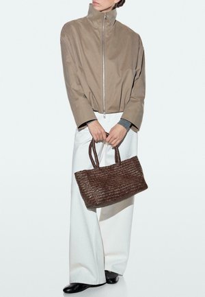 Personne portant une veste beige à fermeture éclair et un pantalon blanc à jambes larges, tenant un grand sac à main tissé marron avec deux poignées.