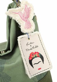 Sac en tissu vert avec un design floral texturé, présentant une étiquette brodée en forme de colibri rose et une étiquette en tissu représentant un visage avec des accents floraux.