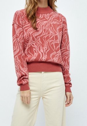 Femme portant un pull à manches longues rose à motifs et un pantalon crème taille haute, debout devant un fond uni.