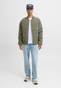 Olijfgroene gewatteerde jas met drukknoopsluiting, witte t-shirt eronder, lichtblauwe recht gesneden jeans en grijze sneakers; marineblauwe pet met logo.