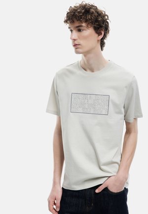 Camiseta de algodón gris claro con cuello redondo, que presenta texto en relieve en gris oscuro. Se combina con jeans de mezclilla oscura.