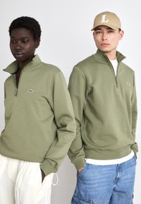 Twee individuen staan naast elkaar in olijfgroene hoodie met rits en een klein logo. De hoodies hebben lange mouwen en een relaxte pasvorm.