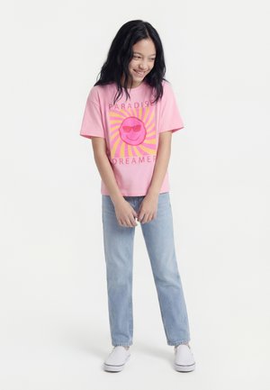 Chica con cabello largo y oscuro, vistiendo una camiseta rosa con la frase "Paradise Dreamer", jeans azul claro y zapatos blancos sin cordones, sonriendo y mirando hacia abajo.