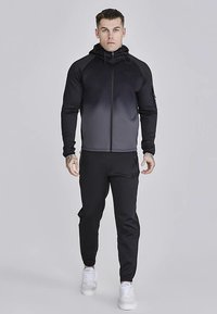 Fekete, elöl cipzáros kapucnis pulóver, karcsúsított szabással, kontrasztos anyagokkal és kenguruzsebbel, fekete, szűk szárú melegítőnadrággal és fehér sportcipővel kombinálva.