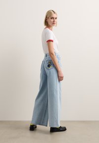Helle blaue, weit geschnittene Jeans mit hoher Taille, die ein gesticktes Design auf der Hintertasche aufweisen. Kombiniert mit einem weißen T-Shirt mit rotem Rand.