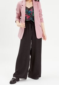 Blazer rose pâle sur un pantalon noir à taille élastique, avec un caraco noir à fleurs colorées orné d'une bordure en dentelle. Chaussures noires épaisses.