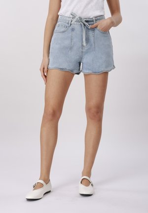 Moteris dėvi šviesiai mėlynas džinsines trumpikes su banguotais kraštais, surištą juosmenį, baltą palaidinę ir baltus Mary Jane batelius, stovinti su viena ranka kišenėje.