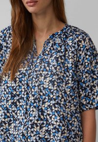 Blauwe en zwarte blouse met patroon, V-hals, korte mouwen en geplooide halslijn. De stof heeft geometrische vormen in verschillende tinten blauw.