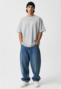 Sivý oversized tričko s krátkymi rukávmi, skombinované s svetlomodrými voľnými džínsami a bielymi teniskami. Čierna čiapka a detail strieborného náhrdelníka.