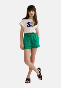 T-shirt en coton avec le texte multicolore "Coucher de soleil", et un short vert avec une taille à cordon, associé à des sandales noires à lanières.