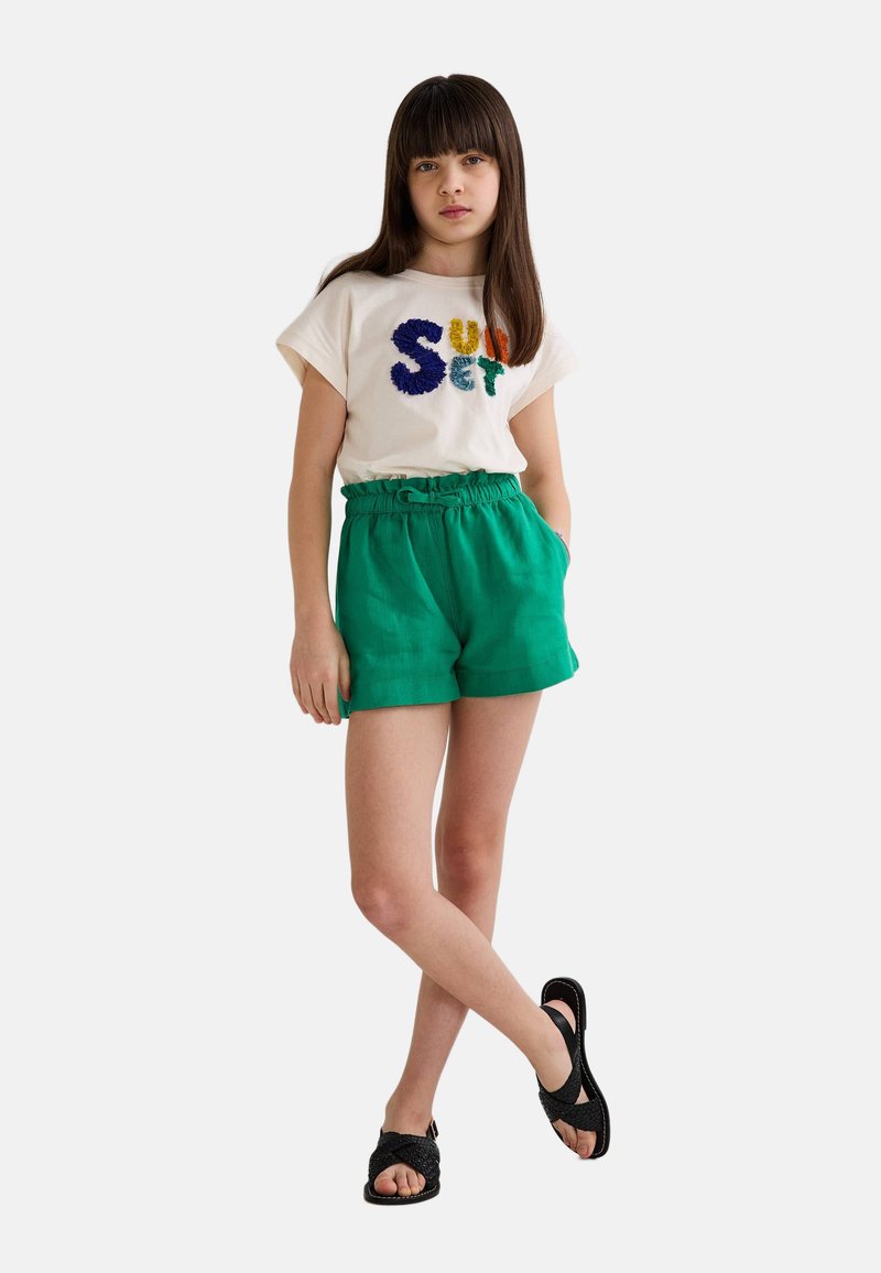 T-shirt en coton avec le texte multicolore "Coucher de soleil", et un short vert avec une taille à cordon, associé à des sandales noires à lanières.
