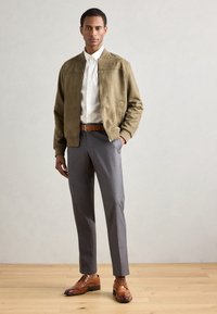 Uomo che indossa una giacca bomber oliva, una camicia bianca, pantaloni grigi, una cintura in pelle marrone e scarpe brogue marroni lucide, in piedi su un pavimento di legno.