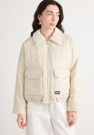 Gequilted beige jas met een kraag, twee voorzakken, ritsluiting en een trekkoord onderaan. Heeft een merketiket aan de onderkant aan de linkerzijde.