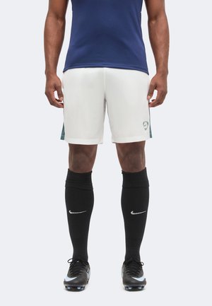 Mandlig atlet iført hvide sportsshorts, sorte knælange Nike-strømper, sorte fodboldstøvler og en mørkeblå trøje, stående med afslappede hænder.
