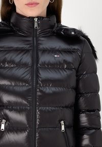 Black puffer jacket with a shiny finish, featuring a high collar, zip closure, and two zippered hand pockets. Fur trim on the hood.