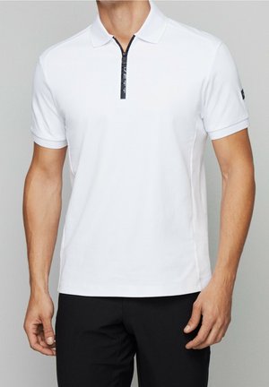 Mann trägt ein weißes Polo mit kurzen Ärmeln und einem schwarzen Reißverschluss vorne, kombiniert mit schwarzen Hosen.