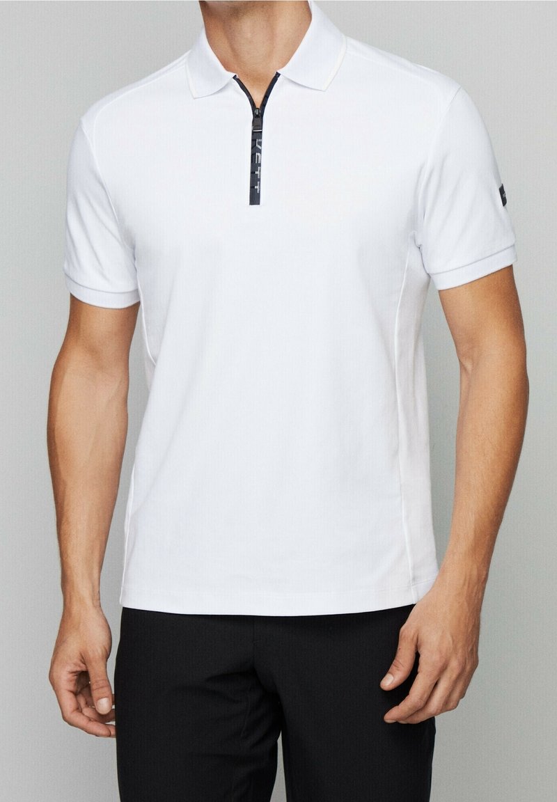 Homme portant un polo blanc à manches courtes avec une fermeture éclair noire sur le devant, assorti à un pantalon noir.