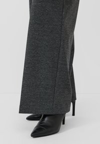 Schwarze High-Heels mit spitzer Schuhspitze, kombiniert mit grauen Wollhosen mit Fischgrätmuster und weitem Schnitt.