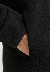 Giacca in pile nera con una superficie morbida e testurizzata; presenta una tasca con zip e polsini a coste. Vista ravvicinata del tessuto e della posizione della mano.