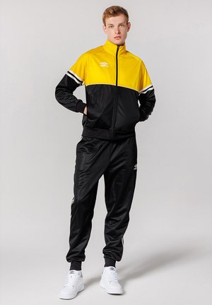 Tuta nera e gialla con giacca zip-up, caratterizzata da strisce bianche e un logo, abbinata a pantaloni coordinati e sneaker bianche.