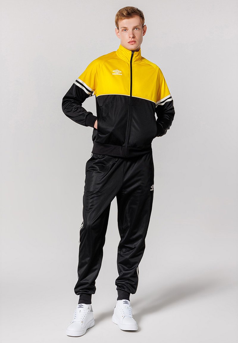 Tuta nera e gialla con giacca zip-up, caratterizzata da strisce bianche e un logo, abbinata a pantaloni coordinati e sneaker bianche.