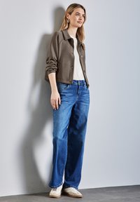 Mörkbrun, kort jacka med knappar över en vit topp, tillsammans med ljusblå, vida jeans och beige sneakers. Neutral bakgrund.