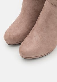 Vaalean taupe-väriset nilkkurit, jotka on valmistettu pehmeästä mokasta. Kenkien muotoilu sisältää pyöreän kärjen ja sileän pinnan, jossa on hieno kiilto ja vähäiset ompelut yksityiskohdat.