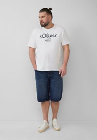 Weißes T-Shirt mit schwarzem Drucktext, kombiniert mit blauen Jeansshorts und weißen Sneakers mit beiger Sohle. Einfaches und lässiges Design.