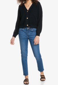 Mujer vestida con un cárdigan negro abotonado, jeans azules y sandalias de estampado de leopardo, de pie contra un fondo blanco.