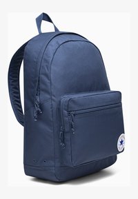 Sac à dos bleu marine en tissu avec une poche frontale zippée, des bretelles rembourrées et un patch logo circulaire. Surface lisse, forme rectangulaire.