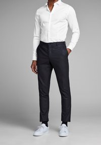 Camicia bianca a maniche lunghe con bottoni, pantaloni scuri su misura e sneakers bianche. Tessuto liscio, vestibilità classica, con dettagli sottili sui pantaloni.