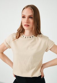 Camiseta de manga corta beige hecha de tela suave, con un cuello redondo y letras negras. Diseño simple con mangas enrolladas.